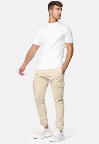 Mladý muž v bílém tričku s krátkým rukávem, béžových cargo kalhotách a bílých teniskách, stojící s jednou rukou v kapse a dívající se na stranu.