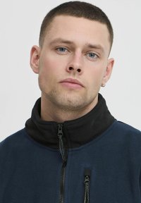 Blue fleece jacket with a black high collar, featuring zippered front pockets and a textured surface. Close-up of the model's face.