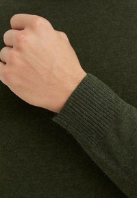 Manche de pull en maille verte avec un poignet côtelé, mettant en valeur une texture lisse et un design ajusté. Main partiellement visible au premier plan.