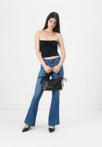 Top negro sin mangas, jeans acampanados azules, sosteniendo un pequeño bolso negro con un acabado texturizado y lazos decorativos, combinado con zapatos negros de punta.