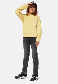 Sudadera amarilla con el logo "Hugo" en relieve, combinada con jeans gris oscuro y zapatillas negras con detalles blancos. Gorra negra.