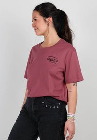 T-shirt en coton bordeaux à manches courtes, avec un logo noir 'OXBOW' sur la poitrine. Associée à un jean noir taille haute.