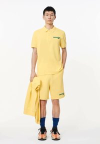 Conjunto de polo amarillo y pantalones cortos con logo verde de "Lacoste". El modelo sostiene un sudadera amarilla a juego. Lleva calcetines azules y zapatillas multicolores.