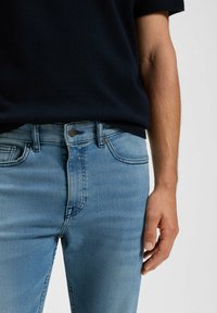 Jeans in denim blu chiaro con vestibilità slim, chiusura a bottone e leggera sbiaditura. Mostrati con una maglietta blu scuro, che mette in risalto la texture dei jeans.