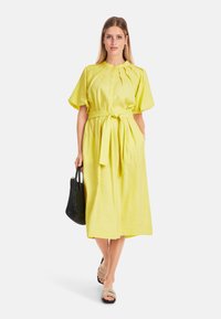 Gelbes Leinen-Midikleid mit kurzen Puffärmeln, vorderen Falten und einem selbstbindenden Gürtel. Das Modell hält eine schwarze geflochtene Tragetasche und trägt beigefarbene Sandalen.