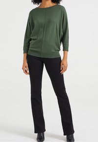 Ciemnozielony, oversize'owy sweter z dzianiny o dekolcie w łódkę i ściągaczami przy mankietach, zestawiony z czarnymi dżinsami w rybaczkach i czarnymi botkami za kostkę.