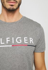 Man med en grå T-shirt med vita bokstäver och ett röd, vit och svart randdetalj över bröstet.