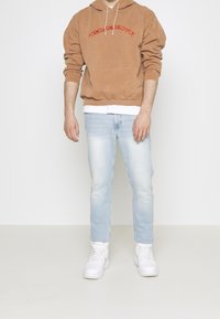 Brown hoodie with red "VINTAGE SUPPLY" embroidery, light blue jeans, and white sneakers. Hoodie features a front pocket and drawstring hood.