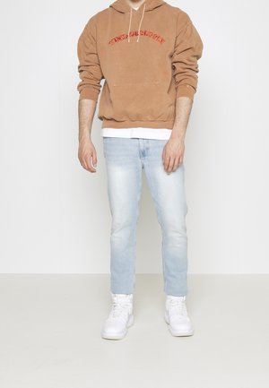 Brązowa bluza z kapturem z czerwonym haftem "VINTAGE SUPPLY", jasnoniebieskie dżinsy i białe sneakersy. Bluza posiada przednią kieszeń i kaptur ze sznurkiem.