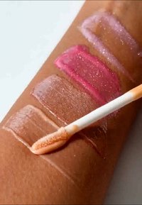 Campioni di lucidalabbra in quattro tonalità: trasparente, nude, rosa e rosa scintillante. È mostrato lo strumento di applicazione, con texture lucide e lisce.