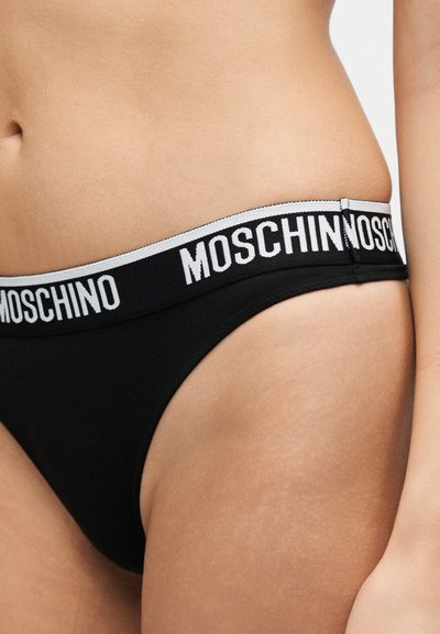 Κοντινό πλάνο του γόνατου ενός ατόμου που φοράει μαύρα εσώρουχα με λευκή ελαστική ζώνη μέσης με το όνομα της μάρκας "MOSCHINO" με έντονα γράμματα.