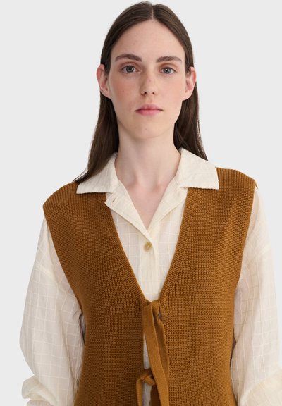 Jeune femme aux longs cheveux bruns portant un chemisier blanc texturé sous un gilet sans manches en tricot marron, tournée vers l'avant sur un fond uni.