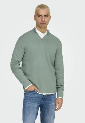 Lichtgroene zip-up trui van zachte stof. Heeft een hoge kraag en lange mouwen, gedragen over een wit shirt en gecombineerd met een blauwe spijkerbroek.