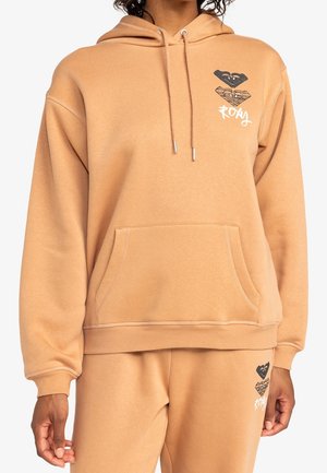 Kobieta ubrana w beżową bluzę z kapturem i spodnie dresowe z czarno-białymi logo w kształcie serc oraz napisem "Roxy" na klatce piersiowej i udzie.