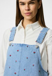 Hele sinine teksariidest üldkleit punaste lillekirjadega, hõbedaste metalldetailide ja reguleeritavate rihmadega, kombineeritud valge kraega särgiga.