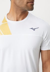 Wit sportshirt met een subtiele gele grafische print op de linkerschouder en een donker logo op de rechterbovenkant. Korte mouwen, ronde hals.