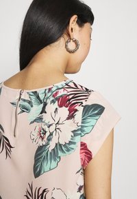 Blouse florale avec un fond rose clair, ornée d'imprimés floraux verts et rouges, manches courtes et un détail de fermeture éclair au dos.