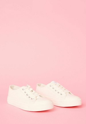 Weiße Leinenschuhe mit einem flachen Design, runden Zehen, sechs metallischen Ösen und strukturierten Gummisohlen. Vor einem pinken Hintergrund.