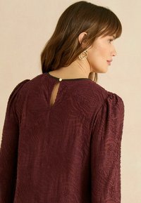 Bordó texturált blúz fodros ujjakkal, hátul kulcslyuk nyílással és fekete szegéllyel. Rétegzett mintás részletekkel és kör alakú arany fülbevalóval.