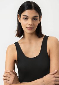 Schwarzes Top mit weitem Ausschnitt und breiten Trägern, aus glattem Stoff gefertigt. Model mit schulterlangen, dunklen Haaren und verschränkten Armen.