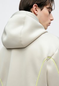 Νεαρό άτομο με κοντά καστανά μαλλιά, φορώντας μια ανοιχτό μπεζ φούτερ με νέον κίτρινες λεπτομέρειες, σε προφίλ μπροστά σε απλό φόντο.