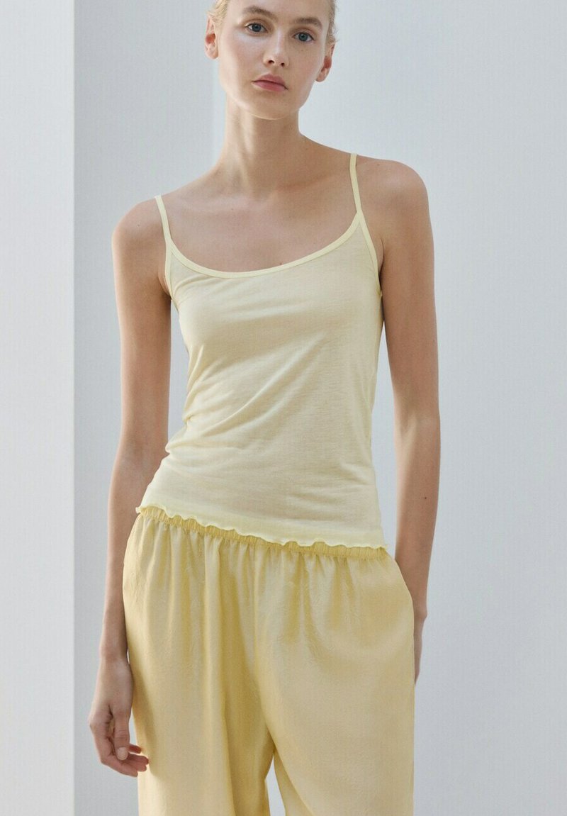 Femme à la peau claire et aux cheveux blonds portant un haut à fines bretelles jaune pâle et un pantalon large jaune sur un fond uni clair.