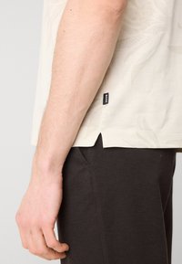 Šviesiai rudos spalvos trumpomis rankovėmis marškiniai su subtiliu lapų raštu, pagaminti iš minkštos medžiagos, su šonine ventiliacija. Suderinti su tamsiai rudais kelnais.