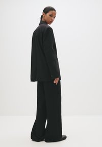 Femeie cu păr împletit în culori închise, îmbrăcată într-un sacou negru lejer și pantaloni negri evazați, întorcându-se peste umăr, pe un fundal alb simplu.