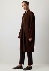 Rudas ilgas kardiganas, minkšta megztinė tekstūra, atviras priekinis dizainas, derinamas su tamsiomis kelnėmis ir loafers tipo batais. Paprasta konstrukcija be papuošimų.