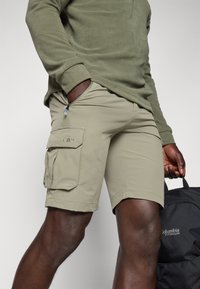 Shorts cargo beiges avec poches latérales et un détail de fermeture éclair, fabriqués en matériau léger, associés à un haut en polaire verte.