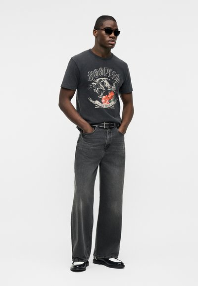 Άνδρας που φοράει μαύρο γραφικό t-shirt με ένα λιοντάρι που βρυχάται και λουλούδια, φαρδιά σκούρα γκρι τζιν παντελόνια, μαύρη ζώνη, γυαλιά ηλίου και μαύρα παπούτσια.