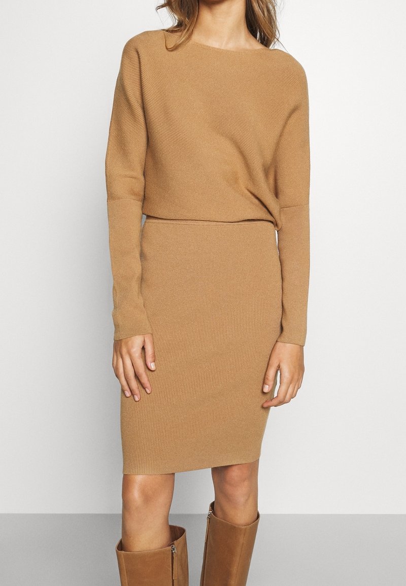 Femme portant une robe en tricot à manches longues couleur chameau et des bottes assorties jusqu'aux genoux, debout devant un fond clair uni.