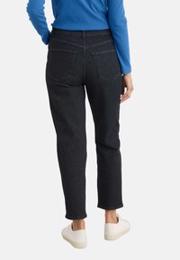 Persoon in zwart hoge taille jeans, blauwe ribben-top met lange mouwen en witte sneakers, gezien vanaf de achterkant tegen een effen achtergrond.