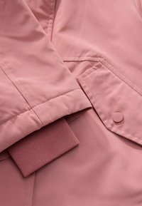 Primer plano de una manga de chaqueta rosa con un puño acanalado y un bolsillo con botón a presión sobre tela lisa.