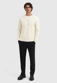 Jongeman met krullend haar, gekleed in een crème crewneck sweater met tekst, zwarte broek en zwarte schoenen, staand met handen in de zakken.