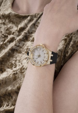 Orologio ottagonale tono oro con accenti di diamanti e cinturino nero testurizzato, indossato su un braccio dalla pelle chiara su uno sfondo di tessuto dorato.