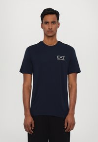 Námořnická modrá tričko s krátkým rukávem vyrobené z měkké látky, s logem na hrudi a černými akcenty na ramenních švech.