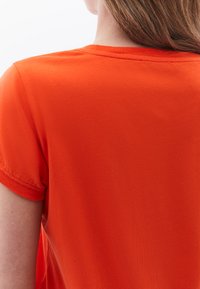 Oranžové tričko s kulatým výstřihem, krátkými rukávy s manžetami a hladkou texturou. Minimální švy kolem výstřihu.