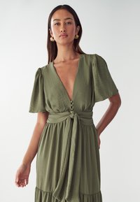Olivengrünes Kleid mit flatternden Ärmeln, tiefem V-Ausschnitt und Knöpfen. Verfügt über eine gebundene Taille und einen fließenden Rock mit gestuftem Saum. Sanfte Textur.