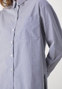Camicia con bottoni caratterizzata da righe verticali blu e bianche, taschino sul petto e vestibilità comoda. Il tessuto appare liscio con una leggera texture.
