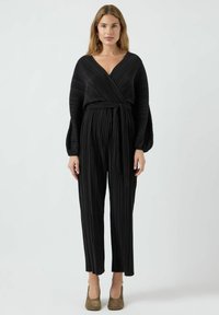 Jumpsuit negro con pliegues, diseño envolvente, mangas largas y cintura ajustada. Presenta un escote en V profundo y piernas rectas.