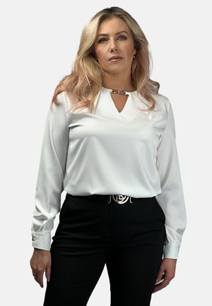 Biała bluzka z długim rękawem z dekoltem w kształcie litery V, z ozdobnym, srebrnym detalem, w połączeniu z czarnymi spodniami i czarnym paskiem.