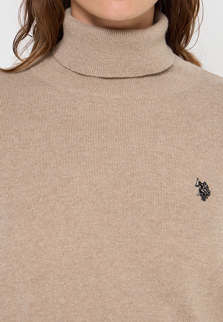 Maglione a collo alto beige realizzato in tessuto lavorato a maglia con texture a coste. Presenta un piccolo logo nero sul lato sinistro del petto.