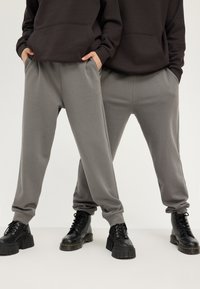 Pantalons de survêtement gris avec des poignets élastiques et des poches latérales, associés à des bottes noires à lacets imposantes, portés par deux mannequins en sweat-shirts à capuche.