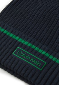 Navy knitted beanie with vertical ribbing, featuring a prominent green horizontal stripe and a rectangular black label with green text.