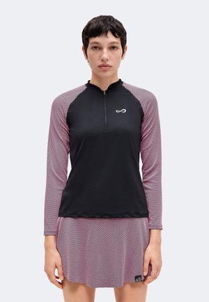 Femme aux cheveux courts et foncés porte un haut noir à manches longues avec des manches roses à motifs et une jupe assortie sur un fond clair uni.