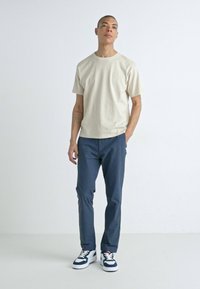 Beige cotton t-shirt with a round neck, paired with dark blue trousers and white sneakers featuring navy accents. Simple, casual outfit.