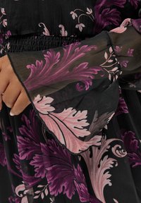 Svart genomskinlig topp i tyg med invecklade rosa och lila blommönster, med smockad midja och klockformade ärmar.