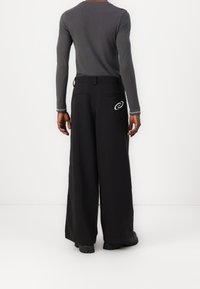 Osoba v černých širokých kalhotách s bílým spirálovým logem na zadní kapse a přiléhavém šedém tričku s dlouhým rukávem, stojící u bílé zdi.