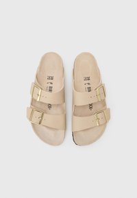 Μπεζ σανδάλια με δύο ρυθμιζόμενους ιμάντες, ορθογώνιες χρυσές αγκράφες και κυρτό σόλα ποδιού. Υφή στη σόλα με αποτύπωμα λογότυπου.